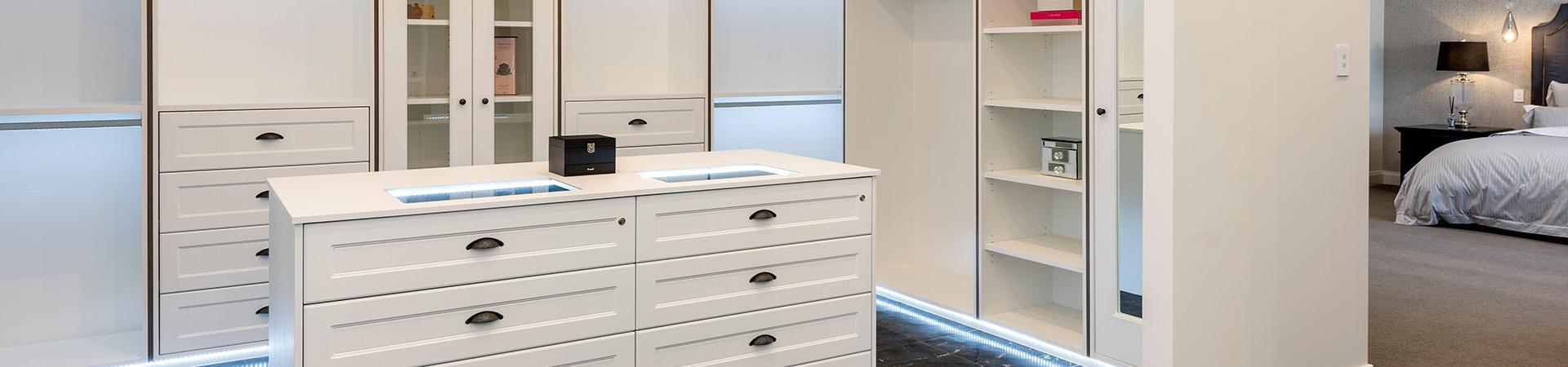Flexi Renovations Perth Custom white walk-in wardrobe with center island drawers, glass-front cabinets, and LED lighting connected to master bedroom