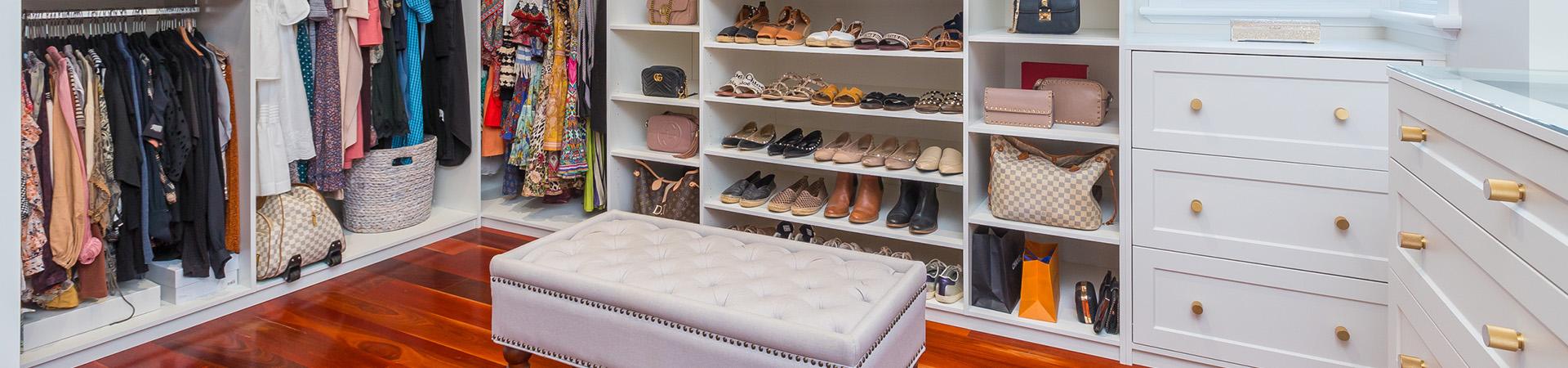 Flexi Renovations Perth Spacious walk-in wardrobe with white custom cabinetry, shoe shelving, hanging clothes, and tufted ottoman on polished timber floor