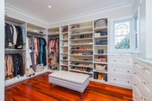 Heritage Elegance: Custom closet with clothes, shoes, bags, ottoman, and wood floor in Nedlands home.